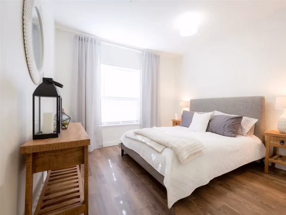 A bright bedroom with a large window covered by light curtains, a gray upholstered bed with white and gray pillows and a white quilt, wooden nightstands with lamps on each side, and a wooden console table with decorative items including a black lantern and a round mirror on the wall.