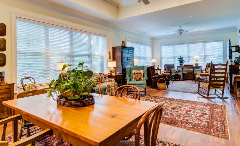 A bright and spacious living room area with large windows covered by white blinds. The room features a wooden dining table with chairs and a green potted plant centerpiece. There are multiple seating options including a green armchair, a striped armchair, a brown leather chair, and a wooden rocking chair. The floor is covered with patterned area rugs, and there are several lamps providing warm lighting. The walls are light-colored, and the ceiling has a fan and recessed lighting.