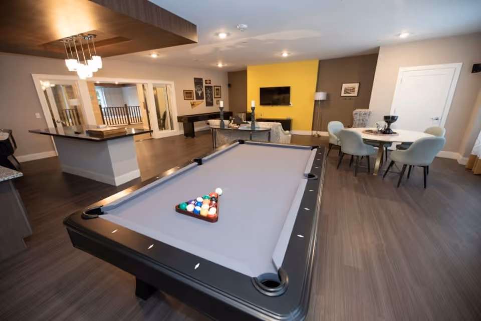 A spacious recreational room featuring a pool table with a rack of billiard balls, a round table with four chairs, a wall-mounted TV on a yellow accent wall, and a seating area with a sofa and a floor lamp. The room has wood flooring, modern lighting fixtures, and a neutral color palette with some decorative wall art.