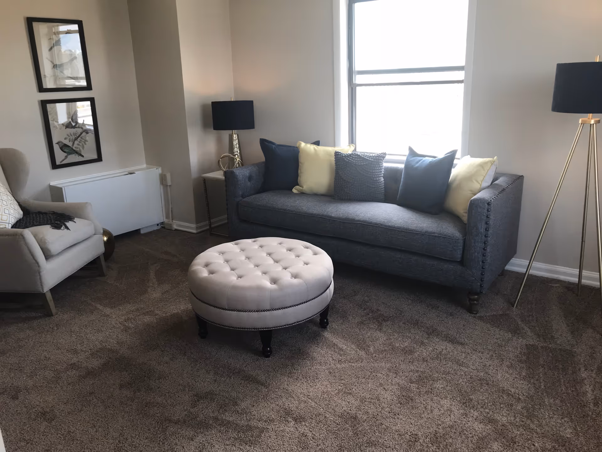 A cozy living room with a gray upholstered sofa adorned with blue and yellow pillows, a round tufted ottoman in front, a beige armchair with a patterned pillow and a dark throw, two framed bird prints on the wall, a small side table with a decorative lamp, and a tall floor lamp near a window letting in natural light.