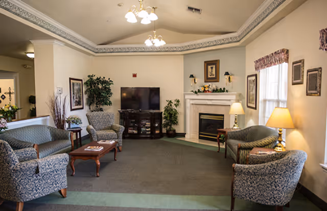 A cozy common living room with upholstered chairs and sofas arranged around a TV and fireplace.