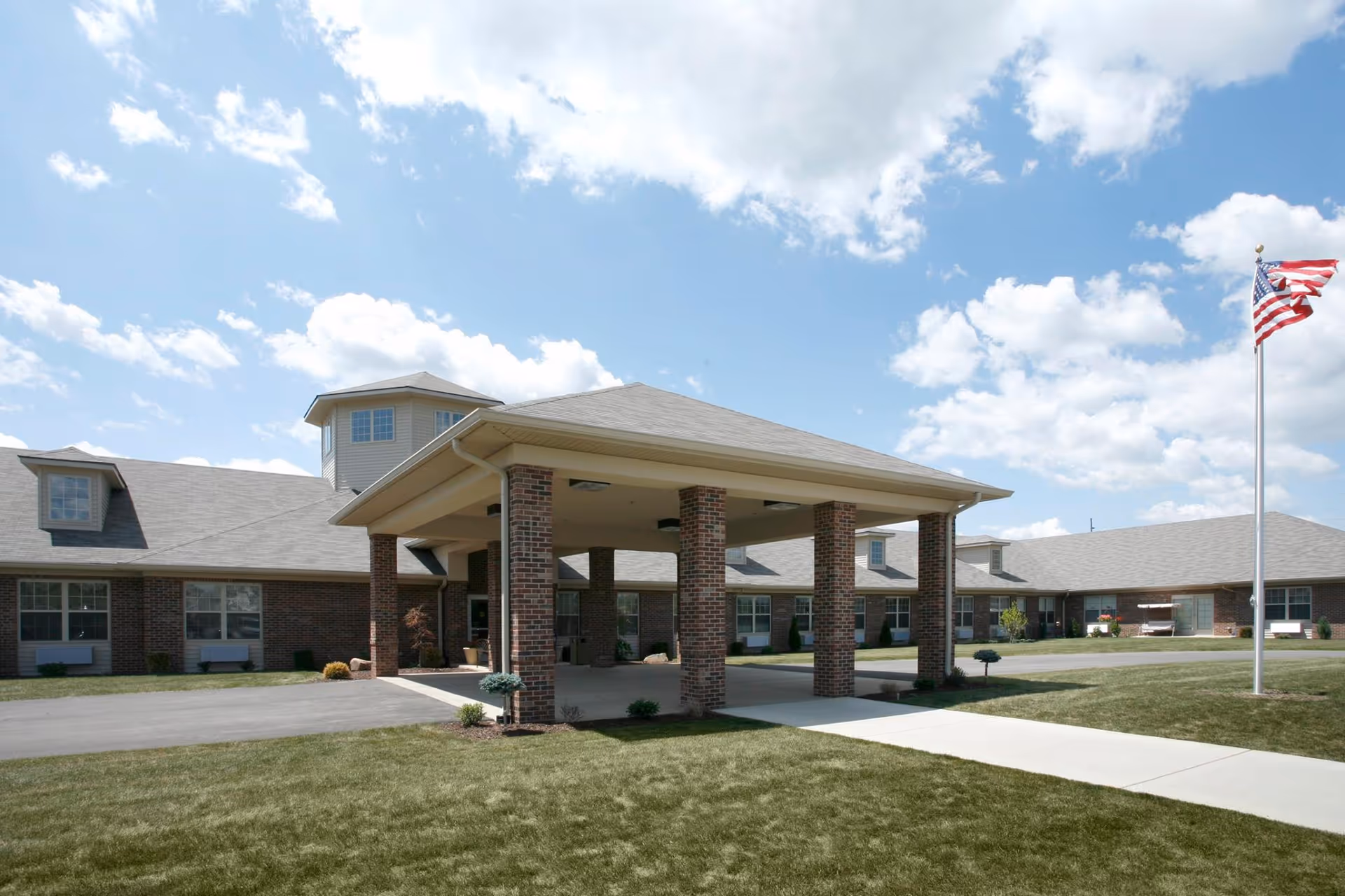 Exterior view of Arbor Trace Family-first Senior Living facility showing a brick building with a covered entrance supported by brick columns, a well-maintained lawn, a sidewalk, and an American flag on a flagpole under a partly cloudy sky.