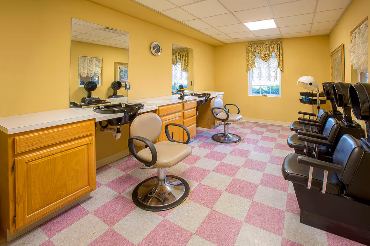 Interior of a hair salon room with yellow walls and a checkered pink and white floor. The room contains two beige salon chairs in front of sinks with mirrors above, and a row of black hair drying chairs with attached hooded dryers along the right wall. A window with decorative curtains is visible on the far wall.