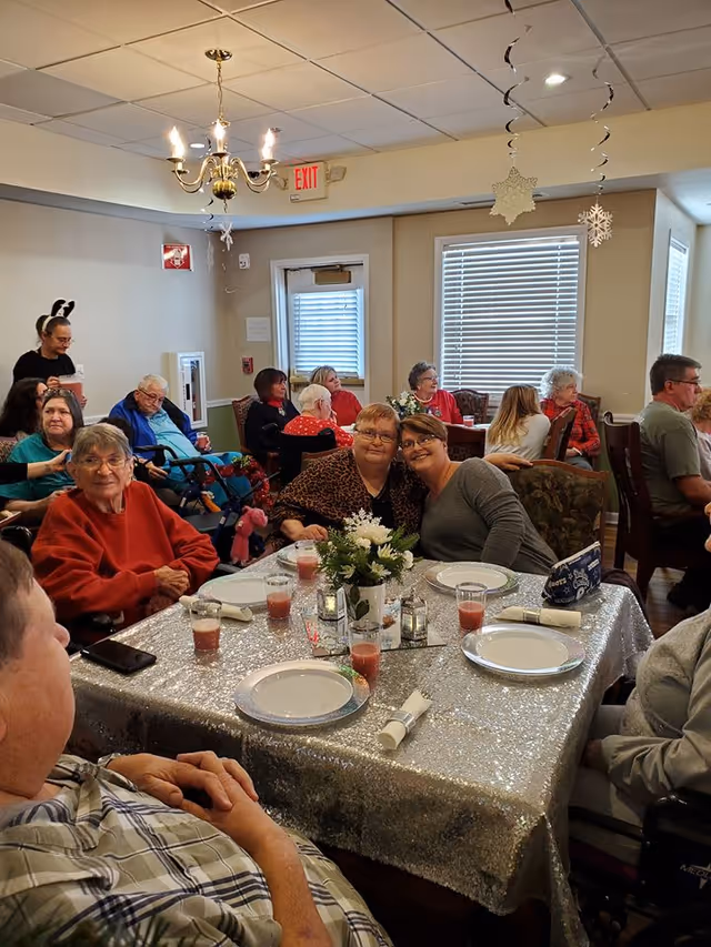 A group of elderly residents seated around a sparkly-decorated dining table in a communal dining room.