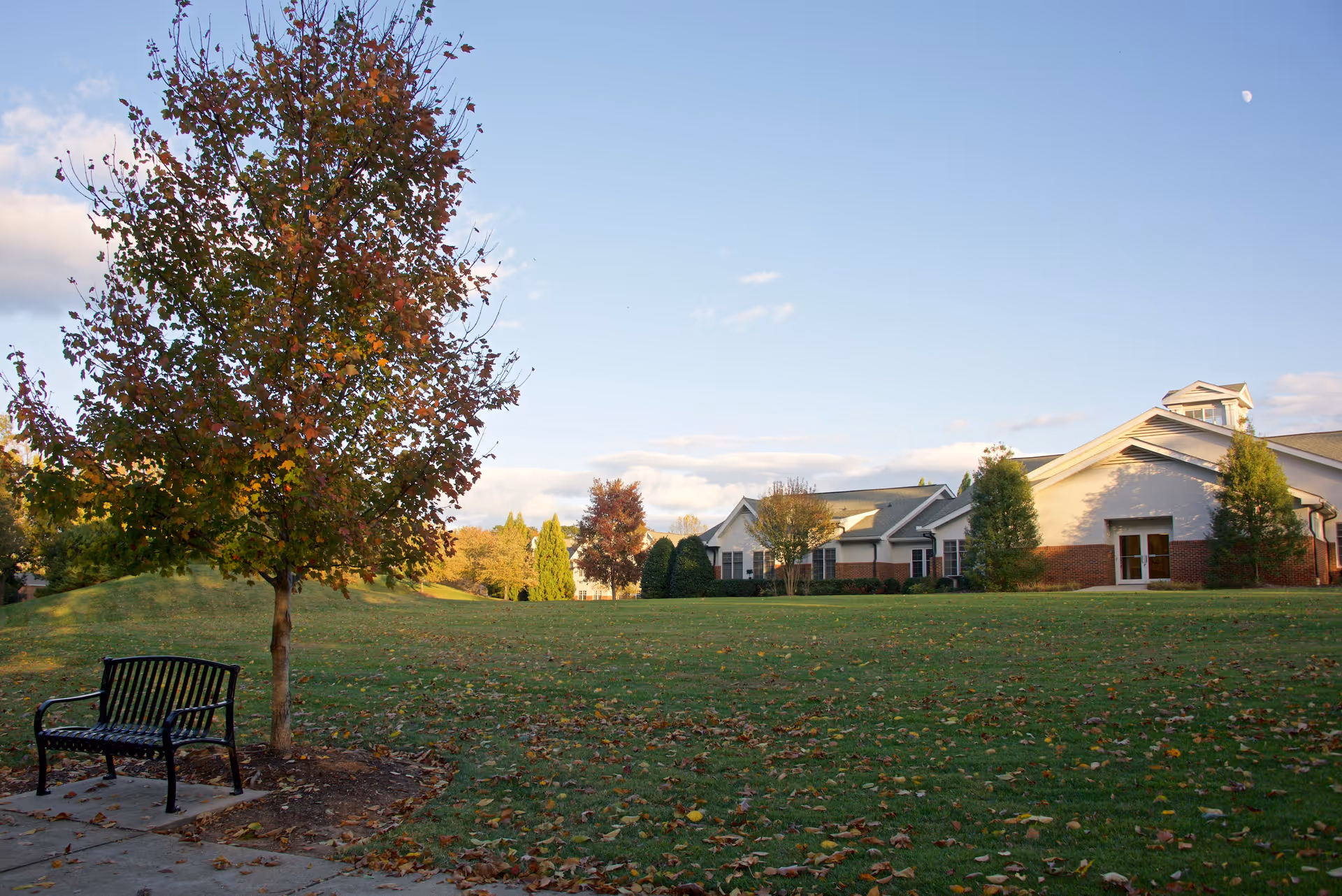 A peaceful outdoor scene at Well-Spring Retirement Community featuring a black metal bench under a tree with autumn-colored leaves, a large grassy area with scattered fallen leaves, and a building with white and brick exterior in the background under a clear blue sky.