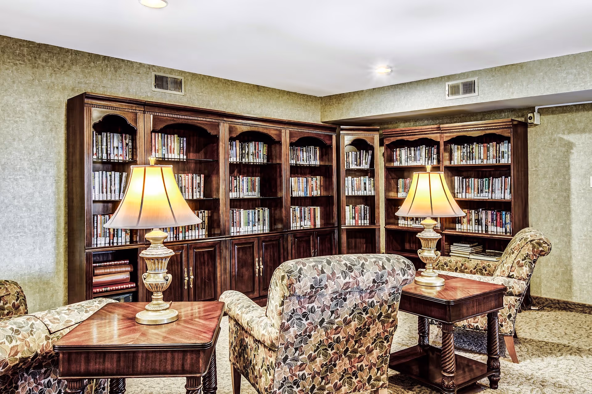 A cozy reading area with floral upholstered armchairs and wooden side tables topped with lamps, set in front of large wooden bookshelves filled with books.