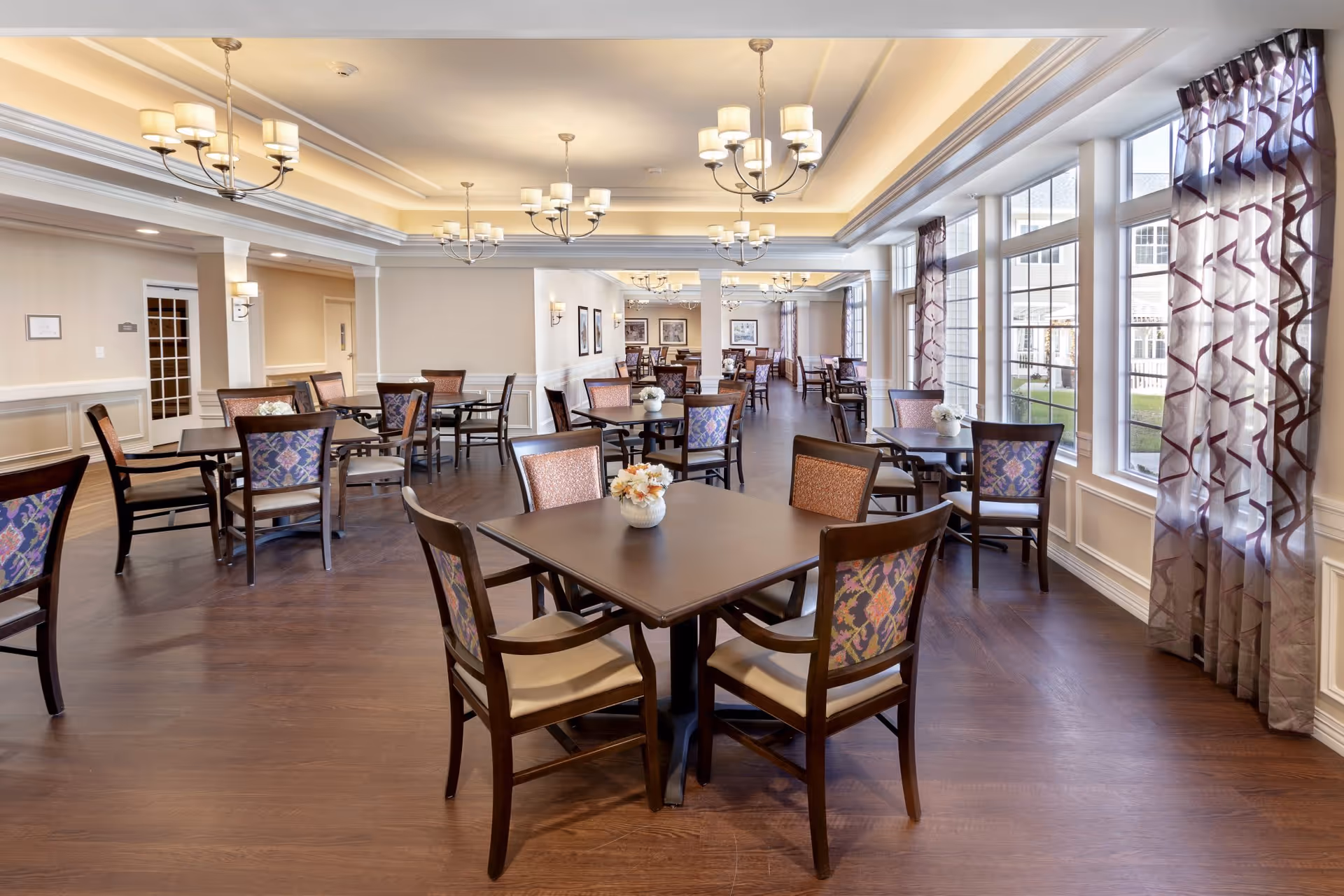 A spacious dining room with multiple square tables and wooden chairs featuring patterned upholstery. The room has large windows with sheer curtains allowing natural light to fill the space. Elegant chandeliers hang from the ceiling, and small floral centerpieces are placed on each table. The floor is wooden, and the walls are painted in a light beige color with white trim.