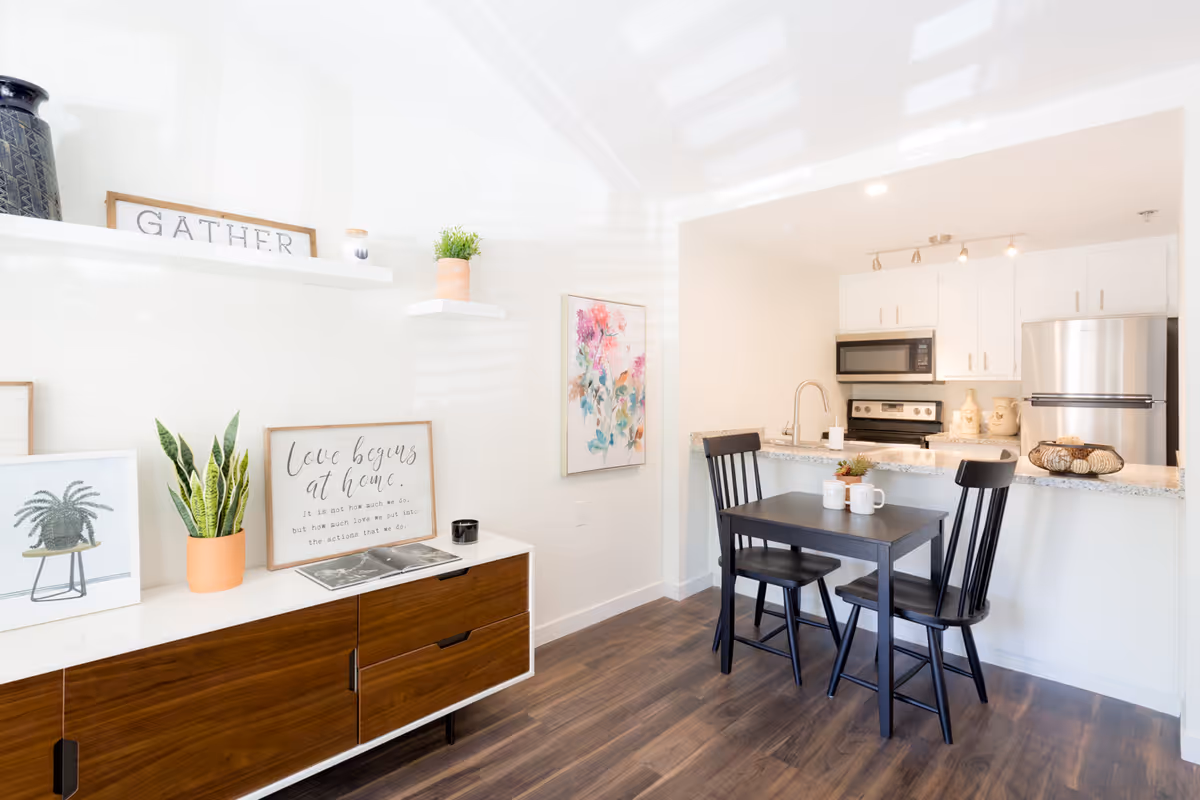 A bright and modern interior space featuring a small dining area with a black table and two black chairs. Behind the dining area is a kitchen with white cabinets, stainless steel appliances including a refrigerator, stove, and microwave, and a granite countertop with a sink. On the left side, there is a wooden sideboard with decorative items including potted plants, framed artwork, and a sign that reads 'Love begins at home.' The walls are white, and the floor is dark wood.