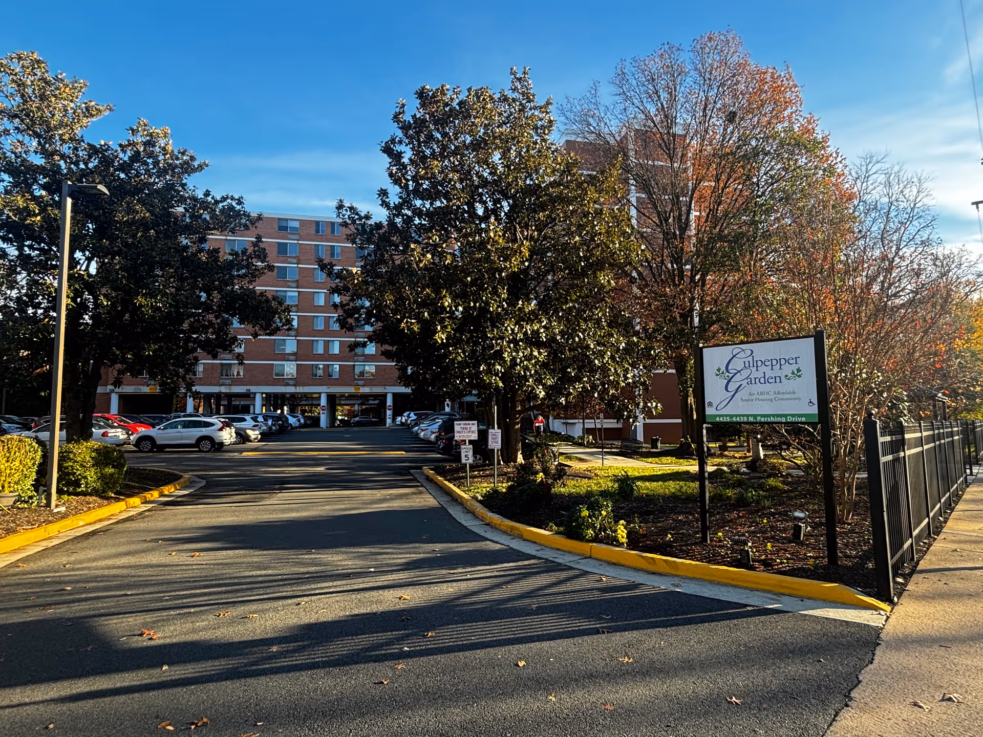 View of the exterior of Culpepper Garden III, a multi-story senior housing building with a parking lot in front, surrounded by trees and landscaping under a clear blue sky.