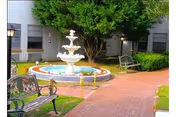 Outdoor courtyard area with a multi-tiered white fountain in the center, surrounded by a circular pool. There are benches and a brick walkway, with green bushes and a large tree providing shade. Windows of the building are visible in the background.