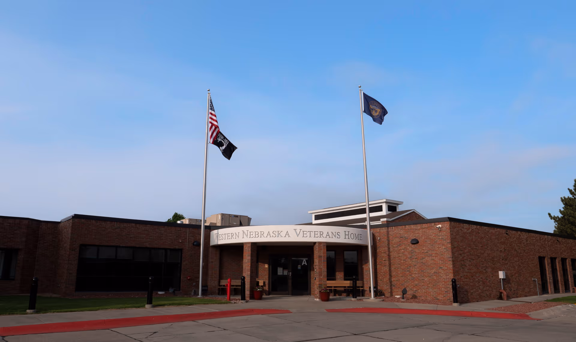 Front exterior view of the Western Nebraska Veterans' Home building with two flagpoles displaying the American flag and other flags against a clear blue sky.