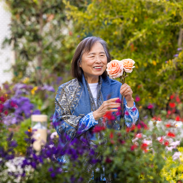 An elderly woman standing in a colorful garden holding two peach-colored roses and smiling, surrounded by various blooming flowers and greenery.
