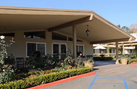 Covered porte-cochere entrance of a nursing center with landscaping, potted plants, outdoor seating, and a driveway.