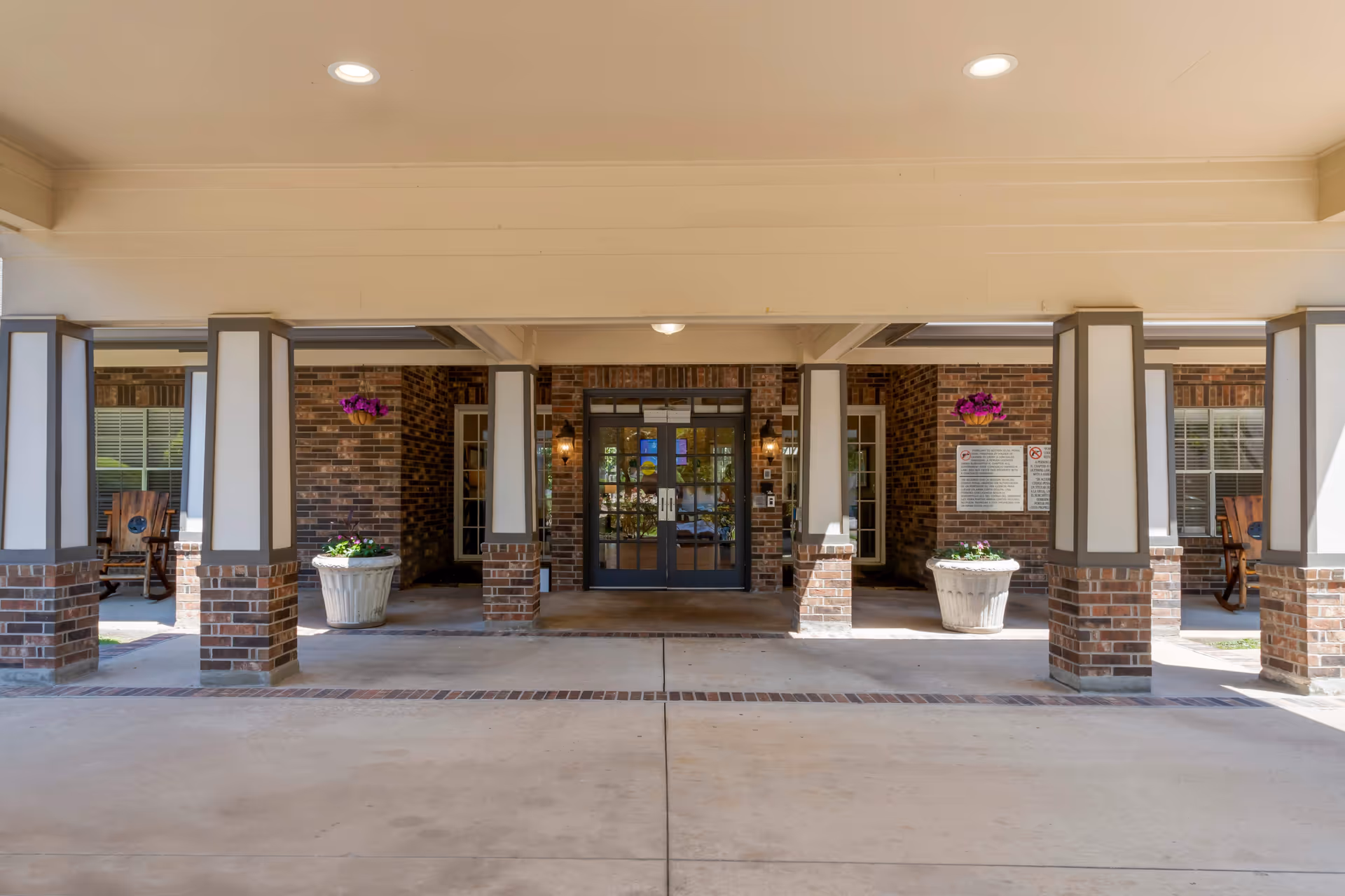 Covered entrance area of a senior living facility with brick pillars and walls, two large white planters with flowers, and double glass doors in the center. There are wooden rocking chairs on either side near windows, and ceiling lights above.