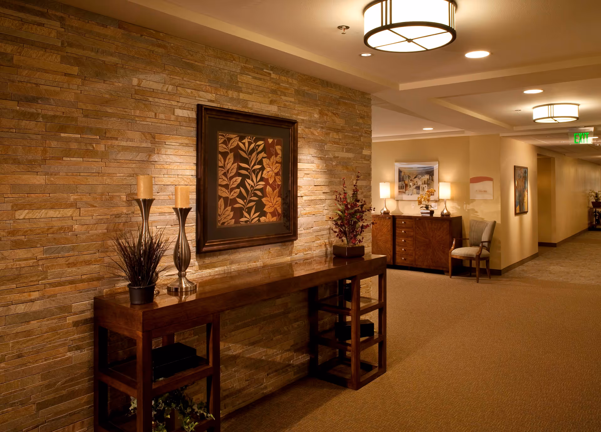 Interior hallway of a senior living facility with a stone accent wall featuring a framed floral artwork. A wooden console table holds two tall candle holders and a potted plant. Further down the hallway, there is a wooden cabinet with two lamps and a chair beside it. The hallway is warmly lit with ceiling lights and has beige walls and carpeted floors.