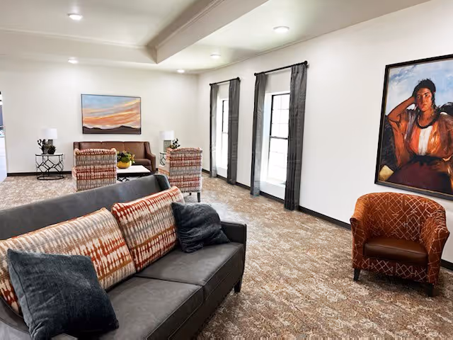 A spacious living room area with a gray sofa adorned with patterned and solid pillows, two patterned armchairs, and a brown armchair. The room has large windows with dark curtains, a carpeted floor, a painting of a woman on the wall, and a landscape painting above a small table with lamps and plants.