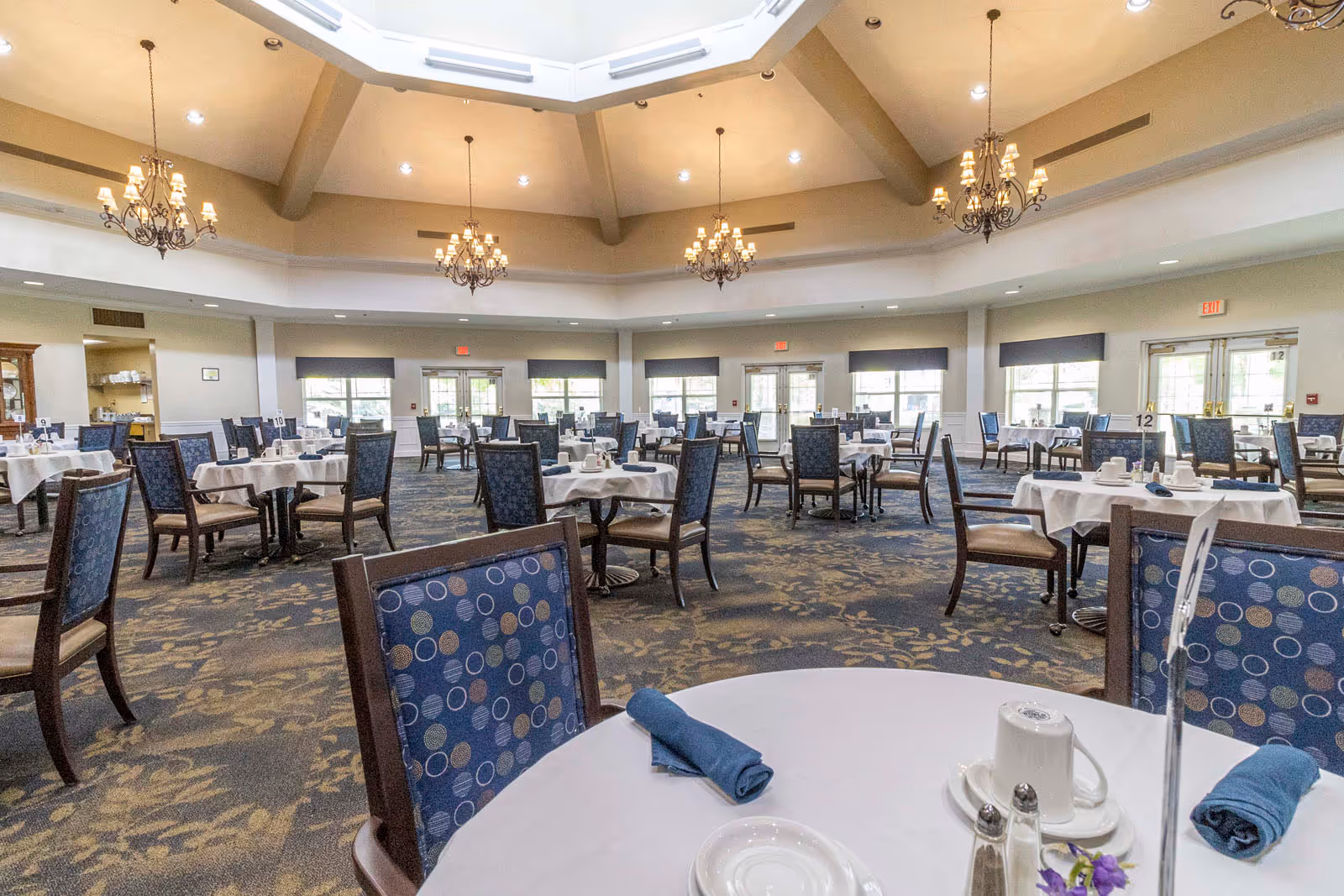 Spacious dining room with multiple round tables covered with white tablecloths, each set with blue napkins, white cups, saucers, and silverware. The room has patterned carpet, large windows letting in natural light, and several ornate chandeliers hanging from a high ceiling with skylights.