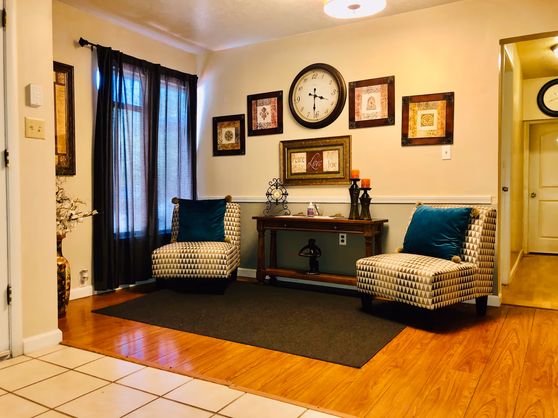 A cozy sitting area in an assisted living care home featuring two patterned armchairs with teal cushions placed on a dark rug. Between the chairs is a wooden console table decorated with candles, a small clock, and a bottle of hand sanitizer. The wall behind the table has a large round clock and several framed decorative art pieces. The floor transitions from tiled to wood, and a window with dark curtains is on the left side.
