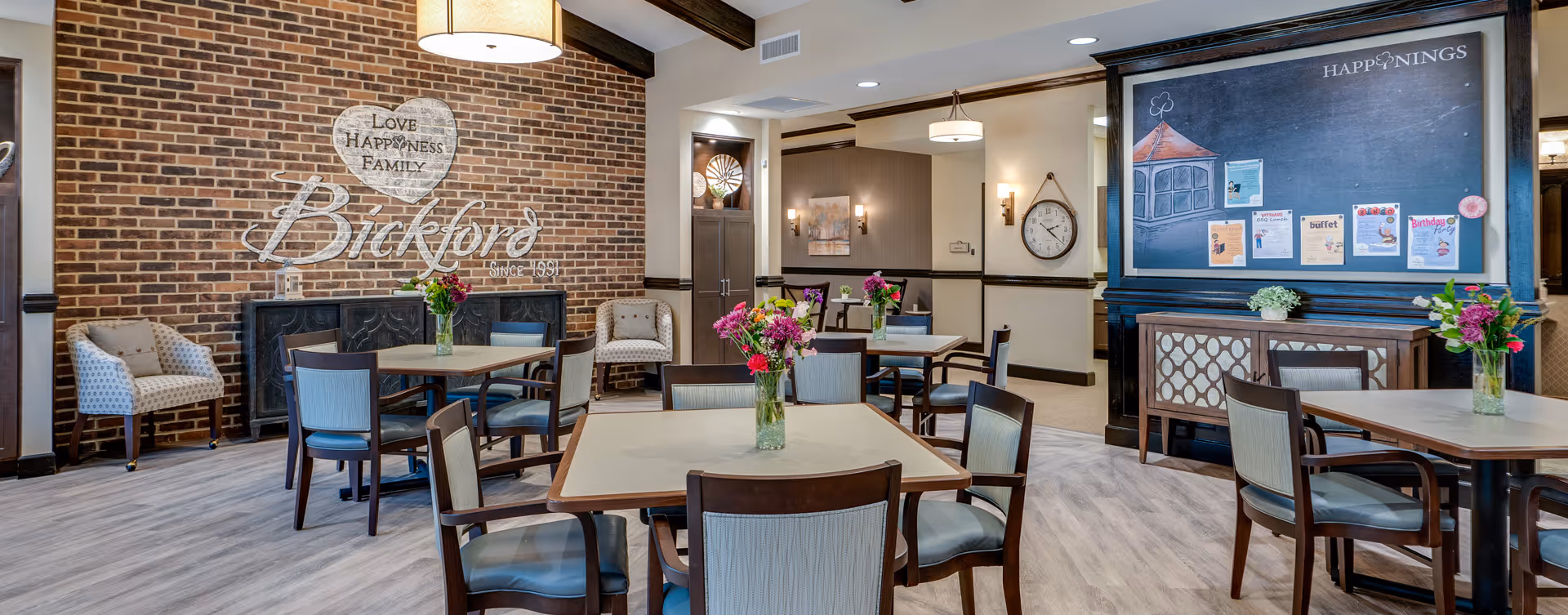 A cozy dining area in a senior living facility with several square tables surrounded by chairs. Each table has a vase with colorful flowers. The room features a brick accent wall with the words 'Love Happiness Family Bickford Since 1991' painted on it. There are two upholstered armchairs near the wall, a large clock on another wall, and a bulletin board labeled 'Happenings' with various flyers and notices. The flooring is light wood, and the ceiling has exposed wooden beams with hanging light fixtures.