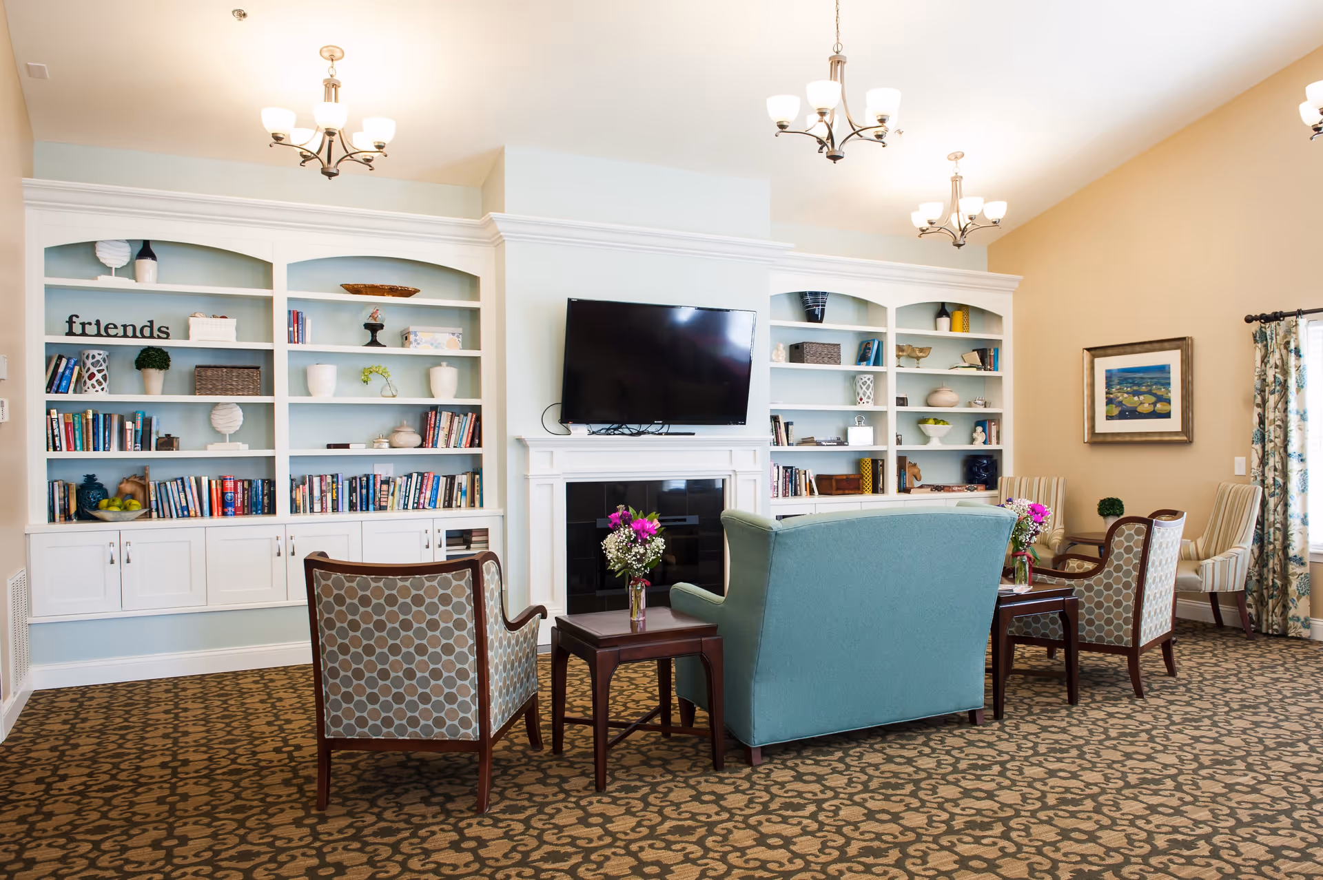 A cozy senior living common area with patterned carpet, a teal loveseat, two patterned armchairs, and a wooden coffee table with a vase of flowers. Behind the seating area is a white built-in bookshelf filled with books and decorative items, and a flat-screen TV mounted above a fireplace. The room is well-lit with ceiling chandeliers and natural light from a window with floral curtains.