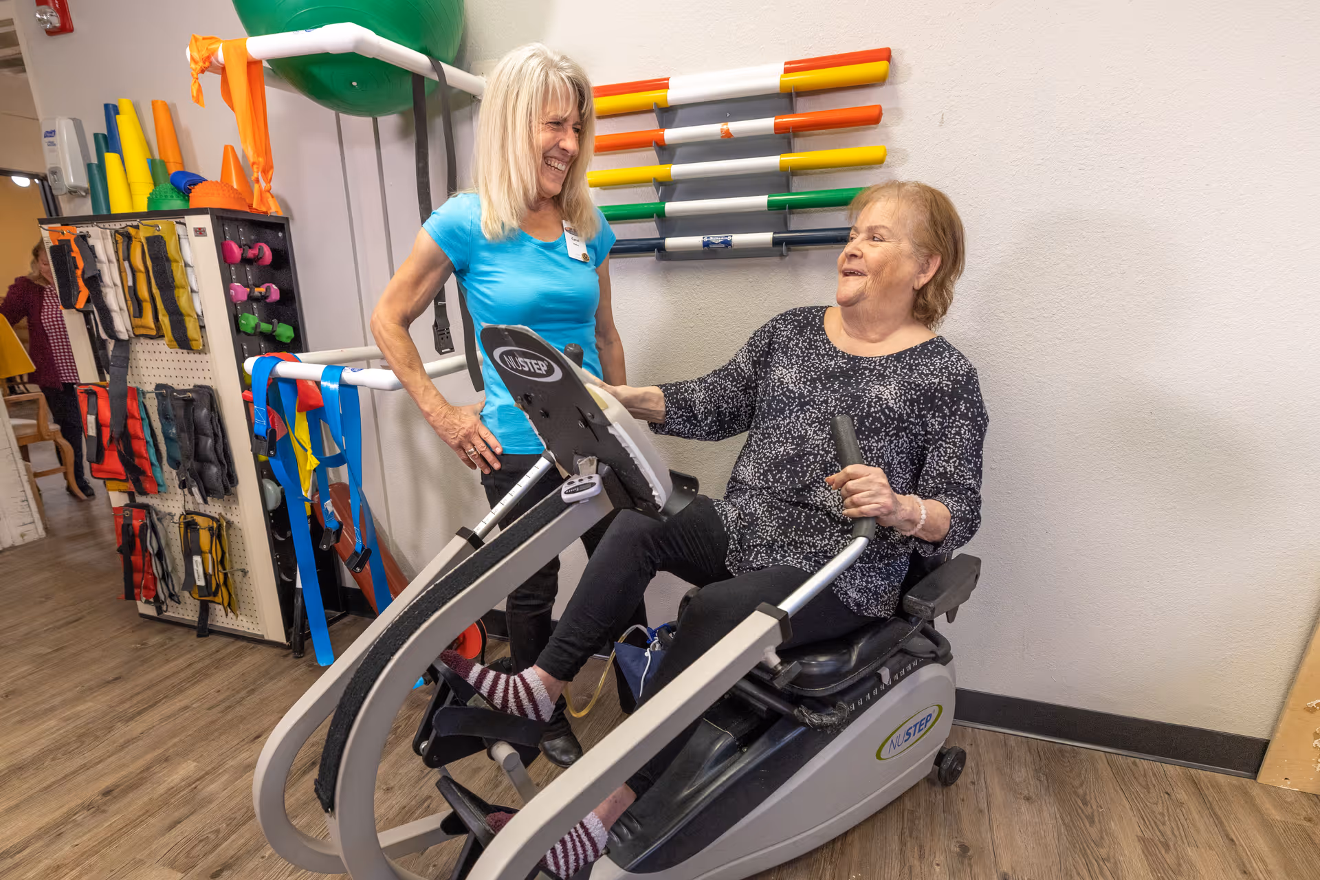 An elderly woman using a seated exercise machine in a rehabilitation or therapy room, smiling and interacting with a female staff member who is standing beside her. The room has various colorful exercise equipment and therapy tools on the walls and shelves.