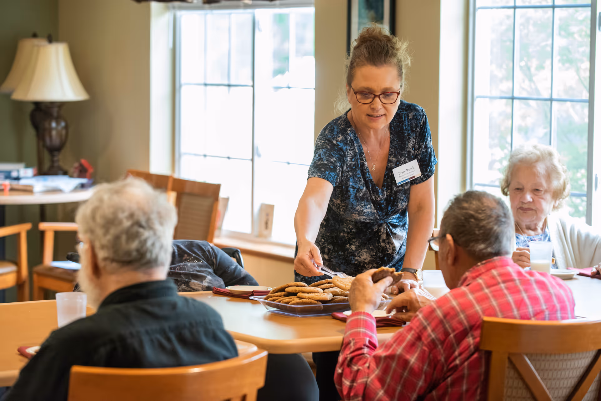 A staff member serves cookies to elderly residents seated around a dining table in a bright common room.