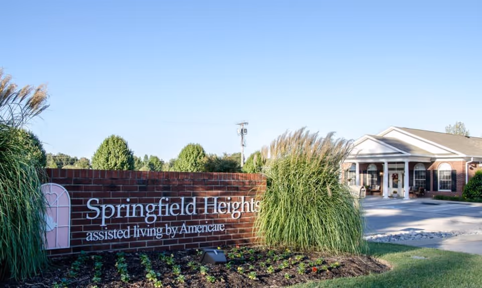 Entrance to Springfield Heights assisted living facility with a brick sign displaying the facility name and landscaping with tall grasses and small plants. The building with a white columned porch is visible in the background under a clear blue sky.