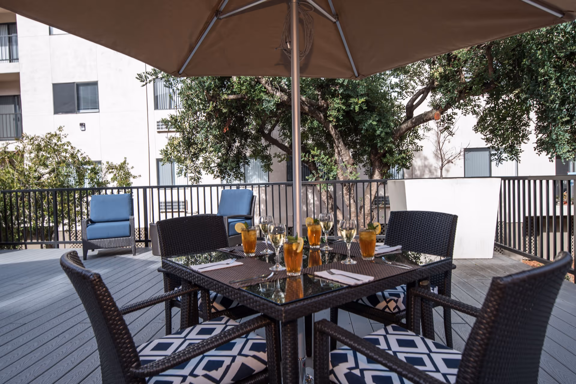 Outdoor patio area with a glass-top table set for four, featuring glasses of iced tea and wine, napkins, and utensils. The table is surrounded by wicker chairs with patterned cushions. There is a large umbrella providing shade, and in the background, there are two blue cushioned lounge chairs, trees, and a building with windows and balconies.