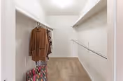 A spacious walk-in closet with beige carpet and white walls. There are two hanging rods on either side, with a few clothing items hanging on the left rod and a colorful bag on the floor beneath them. The closet is well-lit with a ceiling light.