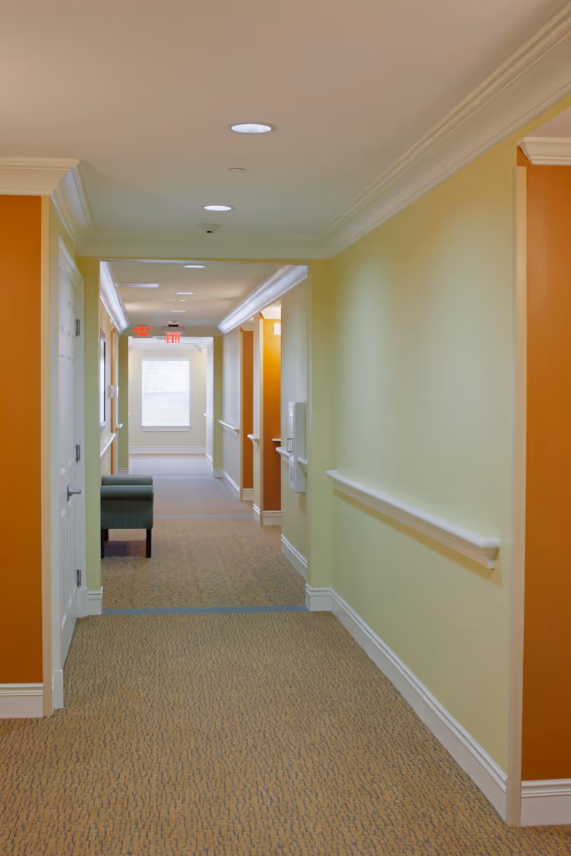 A well-lit hallway in a senior living facility with beige carpet, light yellow and orange walls, white trim, and ceiling lights. There is a green upholstered chair along the left side near a door, and an exit sign is visible at the far end above a window.