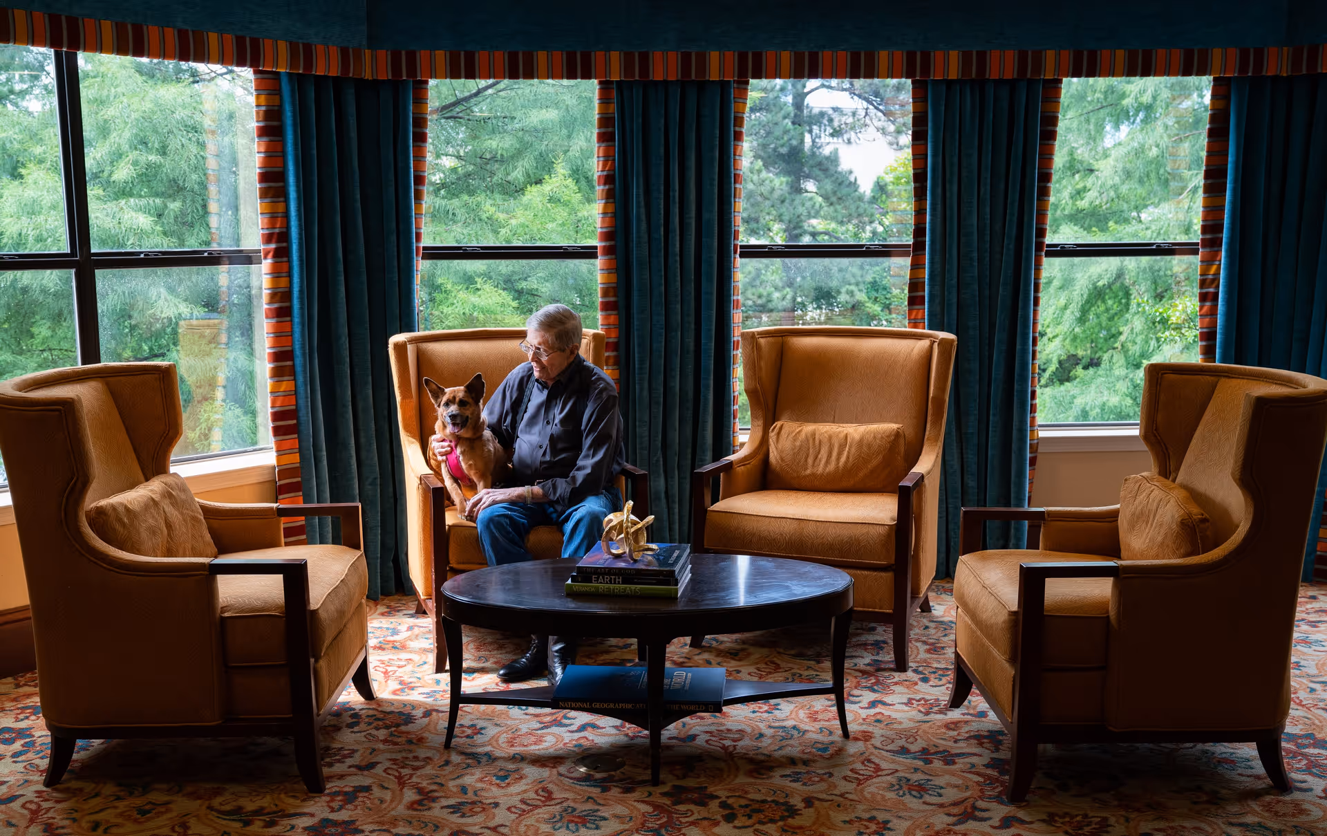 A senior man sitting in a mustard-colored armchair in a cozy living room area, holding a small brown dog wearing a pink harness. The room has large windows with green trees visible outside, dark green curtains with a striped valance, a patterned carpet, and a dark oval coffee table with books and a decorative item on it. Three other matching armchairs surround the coffee table.