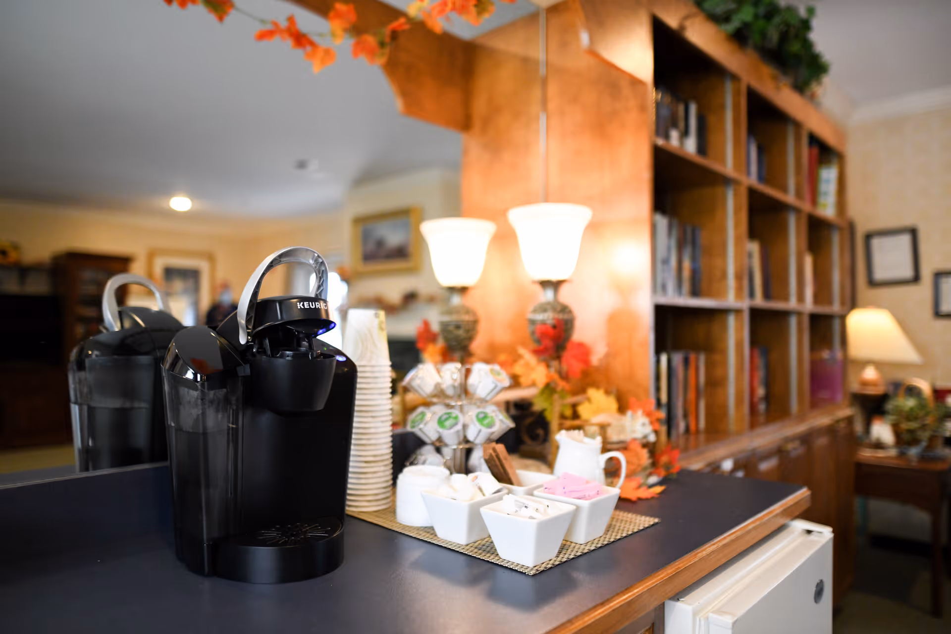 Keurig coffee maker and cups on a countertop in a cozy senior living common room with bookshelves and lamps behind.