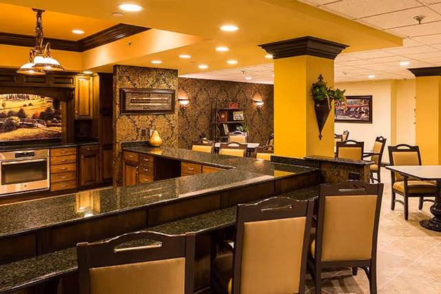 Interior view of a senior living facility kitchen and dining area with dark granite countertops, wooden cabinetry, bar stools, and several dining tables and chairs. The walls are painted yellow and decorated with framed artwork and a wall-mounted plant holder.
