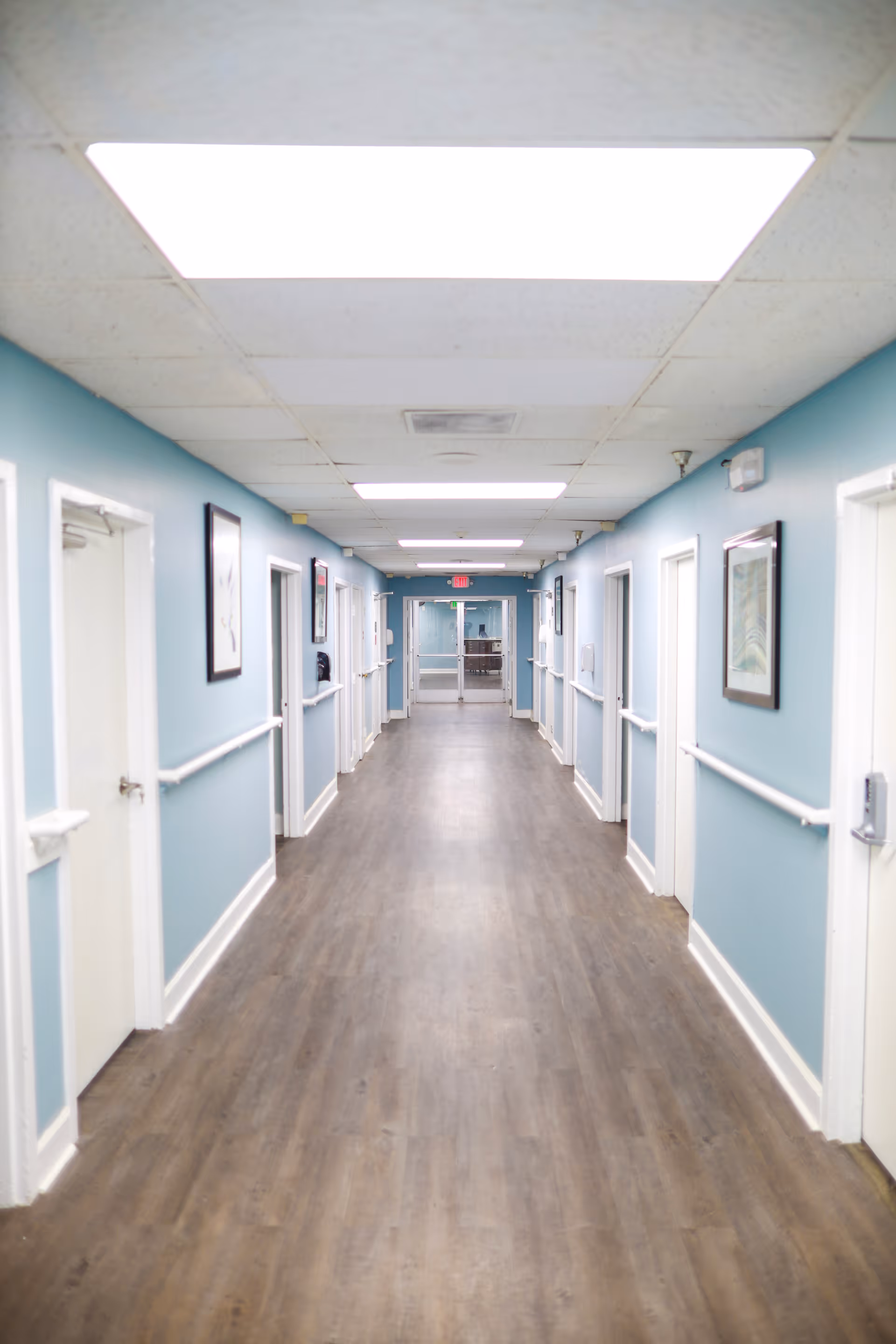 A long, clean hallway in a healthcare facility with light blue walls, white doors on both sides, handrails along the walls, framed pictures, and bright overhead lighting leading to a glass door at the end.