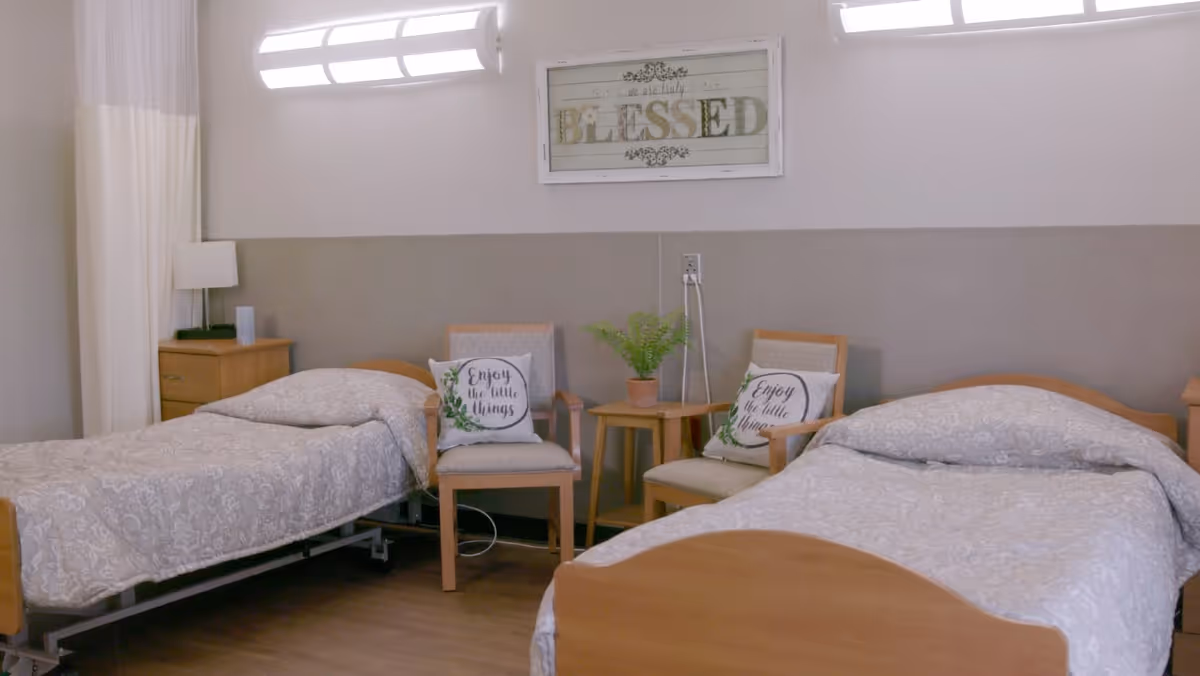 A shared nursing room with two beds, chairs, side tables, and wall art.