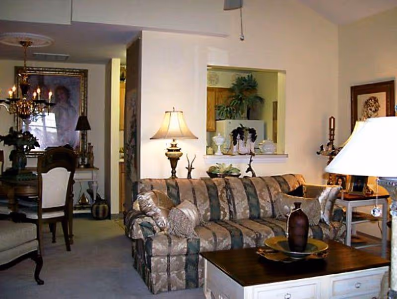 A cozy living room with a patterned sofa, a wooden coffee table with decorative items, two table lamps, and a view into a dining area with a chandelier and framed artwork on the wall.