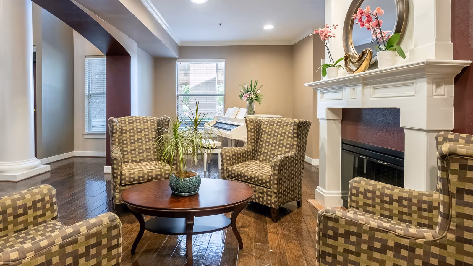 A cozy living room area with four patterned armchairs arranged around a round wooden coffee table with a potted plant. In the background, there is a white piano with a vase of flowers on top, and a white fireplace mantel decorated with pink orchids, a round mirror, and a gold heart-shaped ornament. The room has wooden flooring and neutral-colored walls with large windows letting in natural light.