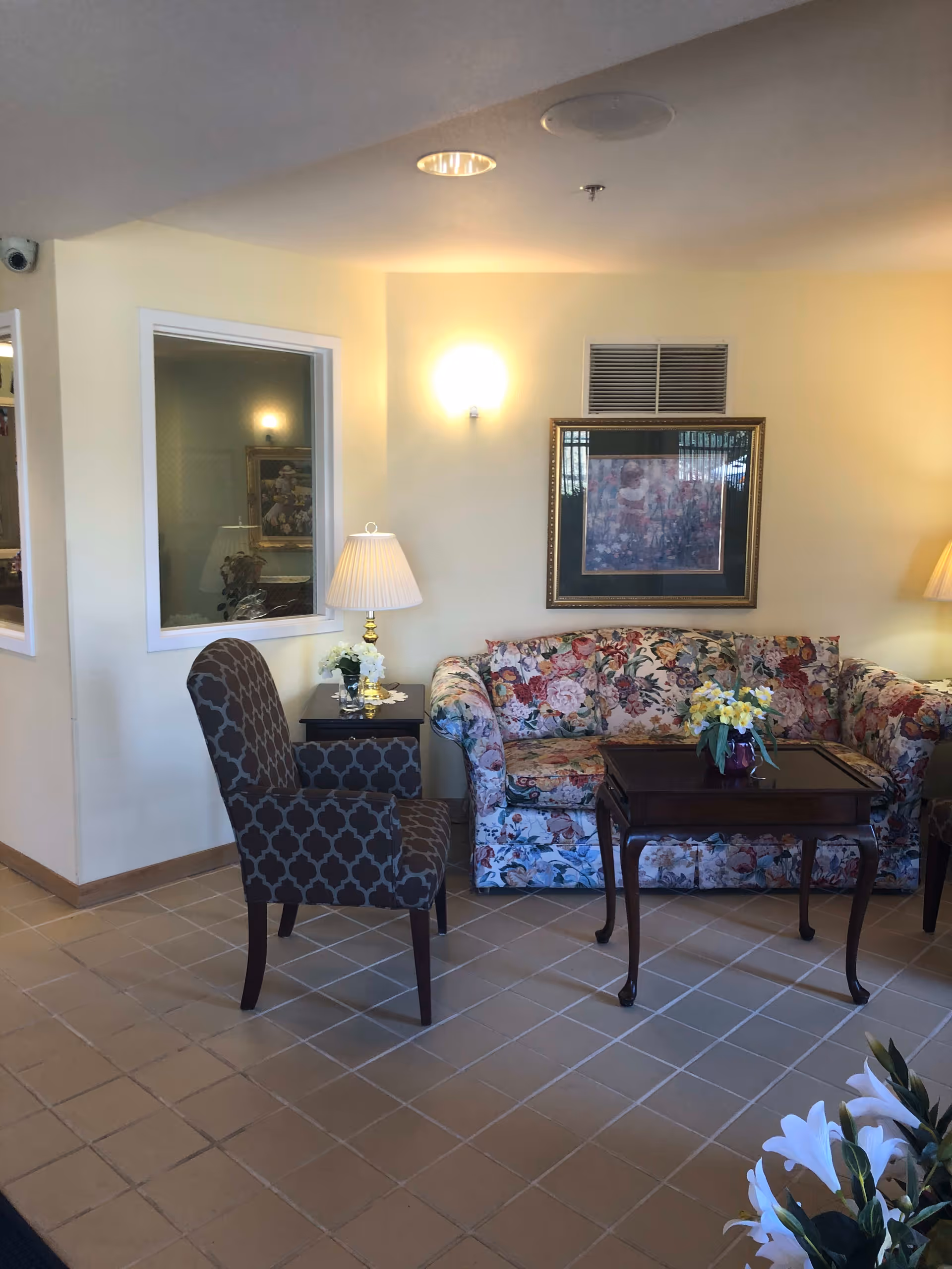 A cozy sitting area in a senior living facility with a floral patterned sofa, a dark wooden coffee table with a small flower arrangement, a patterned armchair, two table lamps on side tables, a framed painting on the wall, and beige tiled flooring.