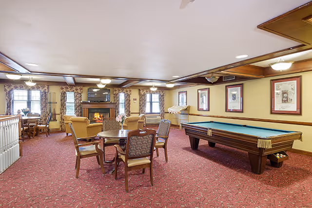 A spacious common room with a red patterned carpet, a pool table, several wooden chairs and tables, and a seating area with armchairs near a fireplace. The walls are decorated with framed pictures, and windows with floral curtains allow natural light into the room.
