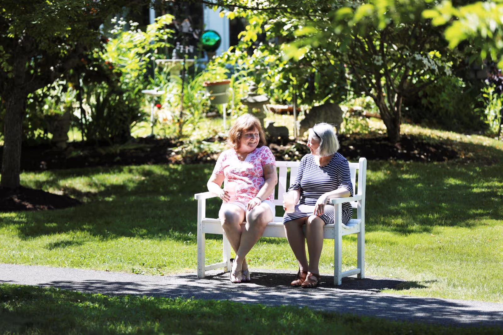 Two older women sitting and talking on a white bench in a sunlit garden.