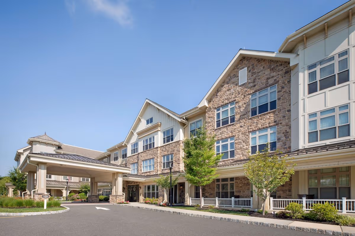 Exterior view of a multi-story senior living facility building with stone and siding facade, large windows, a covered entrance, and landscaped greenery under a clear blue sky.
