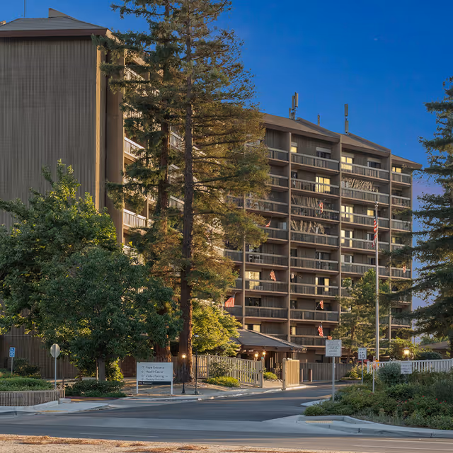 Exterior view of a multi-story retirement community building with balconies, surrounded by tall trees and greenery under a clear blue sky.