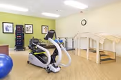 A well-lit physical therapy or exercise room with light green walls and wood flooring. The room contains an elliptical machine, a large blue exercise ball, a rack with various exercise weights, and a wooden staircase with handrails for rehabilitation exercises. Three framed pictures and a clock are mounted on the wall.