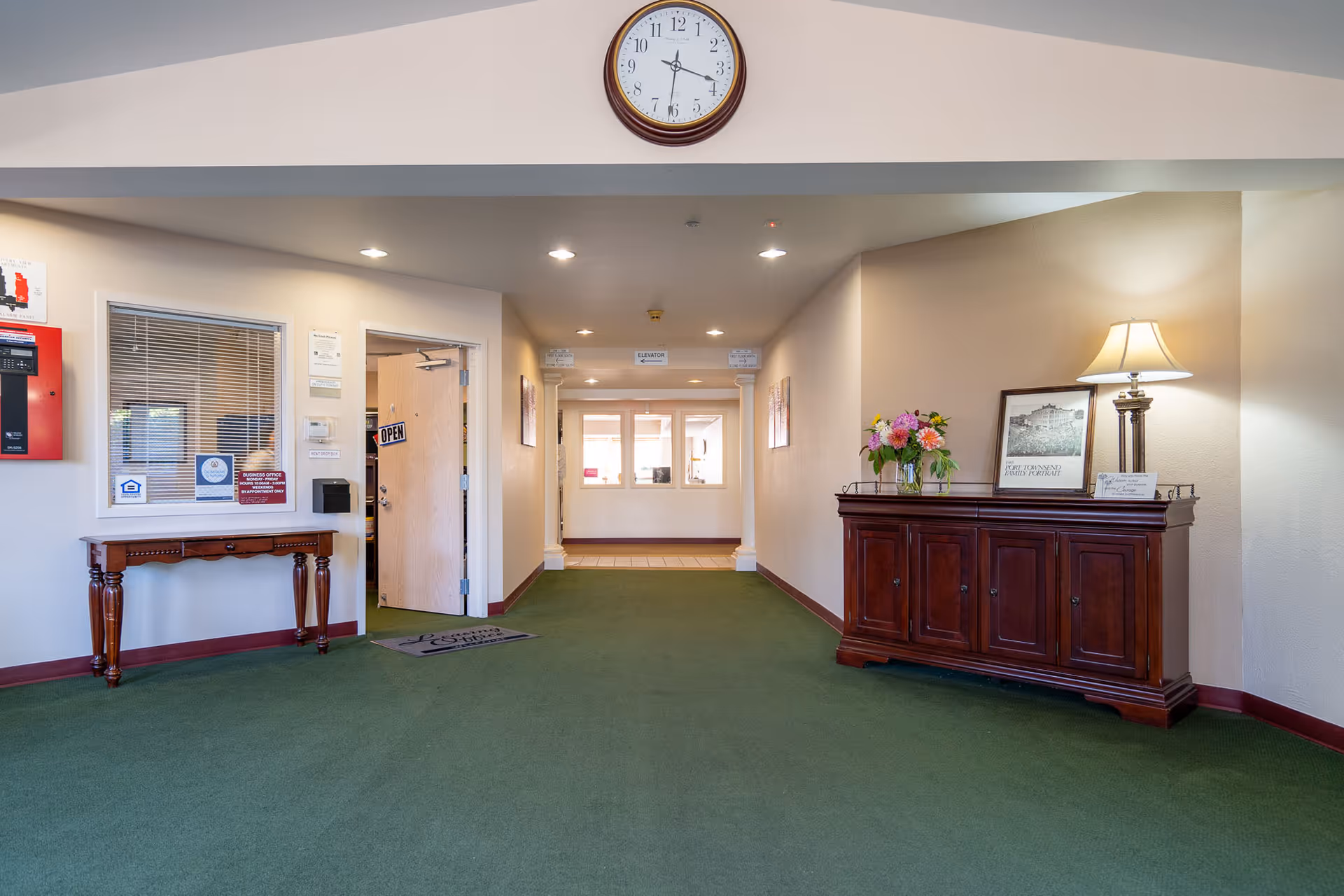Interior hallway of a retirement facility with green carpet, a wooden side table on the left, and a wooden cabinet with a lamp, flowers, and framed pictures on the right. A clock is mounted on the wall above the hallway entrance, and an open door with an 'OPEN' sign is visible on the left side.