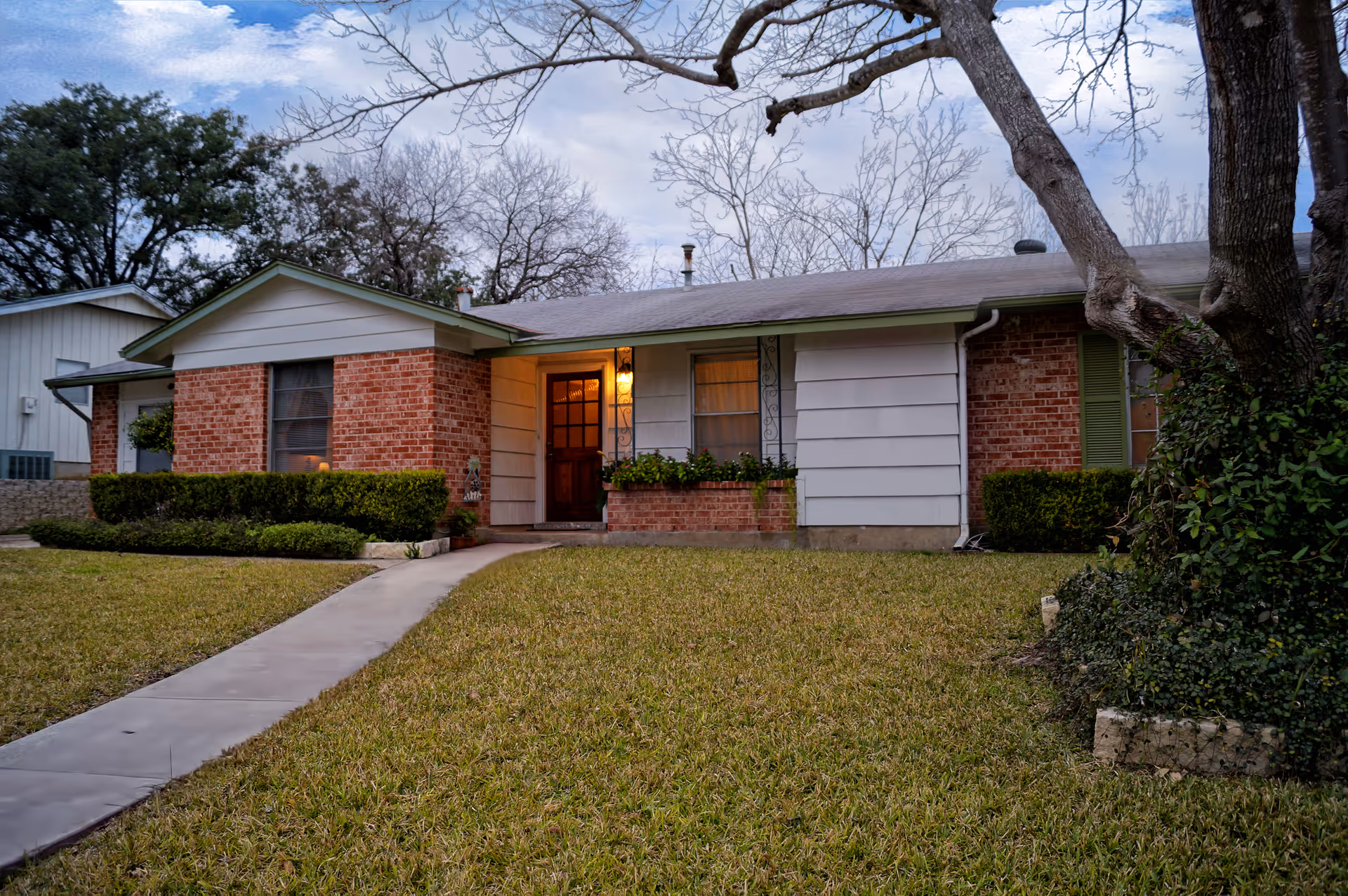 A single-story brick and siding house with a front door illuminated by a porch light. The house has a well-maintained lawn, a concrete walkway leading to the entrance, and some bushes and trees around the property. The sky is partly cloudy.