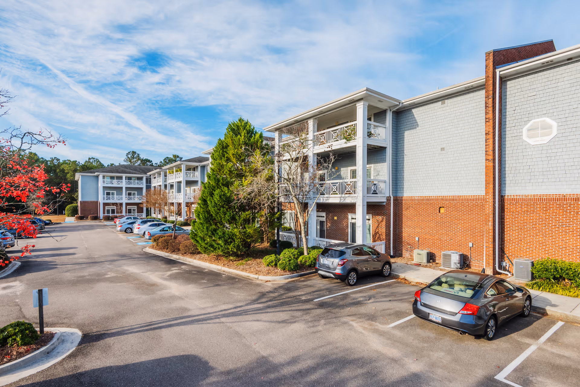 Exterior view of a multi-story senior living building with balconies, landscaping, and parked cars in the lot.