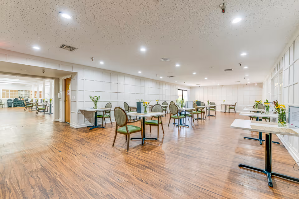 A spacious dining room with wooden flooring and white paneled walls. The room contains multiple small tables, each with two chairs featuring green cushions. Some tables have small flower arrangements. The room is well-lit with recessed ceiling lights and has a clean, organized appearance.