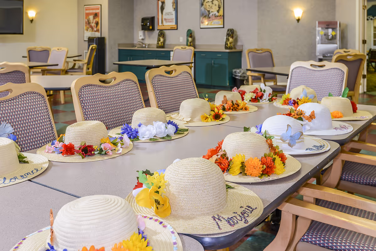 A long table in a communal room with multiple straw hats decorated with colorful artificial flowers and butterflies arranged neatly on top. The room has several chairs with patterned upholstery, a green cabinet with a sink, framed posters on the wall, and a water dispenser in the background.