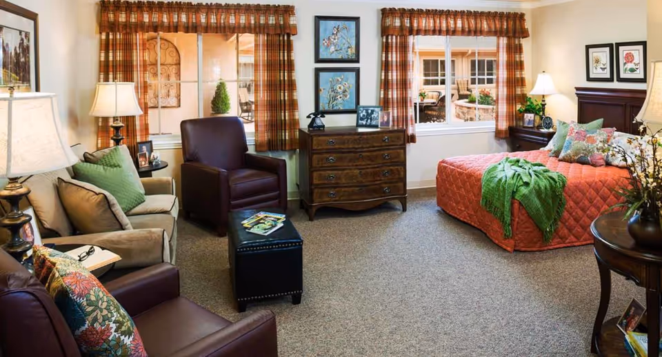 A cozy and well-decorated bedroom featuring a bed with an orange quilt and green throw, two leather armchairs, a beige sofa with green and floral pillows, a wooden dresser, and two windows with plaid curtains letting in natural light. The room also includes several lamps, framed artwork on the walls, and a small round table with a vase of flowers.