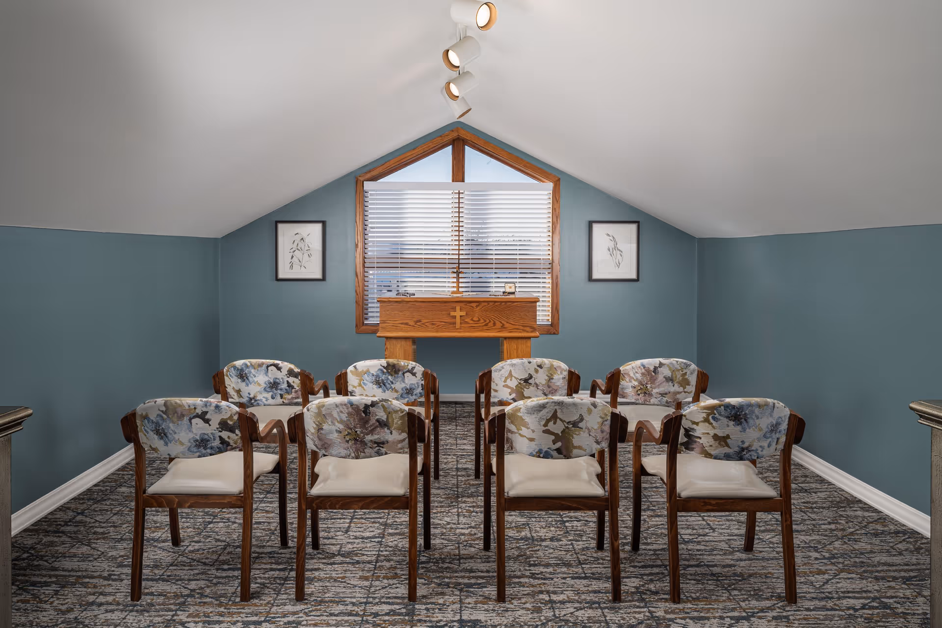 Small chapel-style meeting room with rows of floral-upholstered chairs facing a wooden lectern beneath a triangular window.